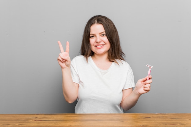 Jeune femme sinueuse de taille plus tenant une lame de rasoir montrant le numéro deux avec les doigts.