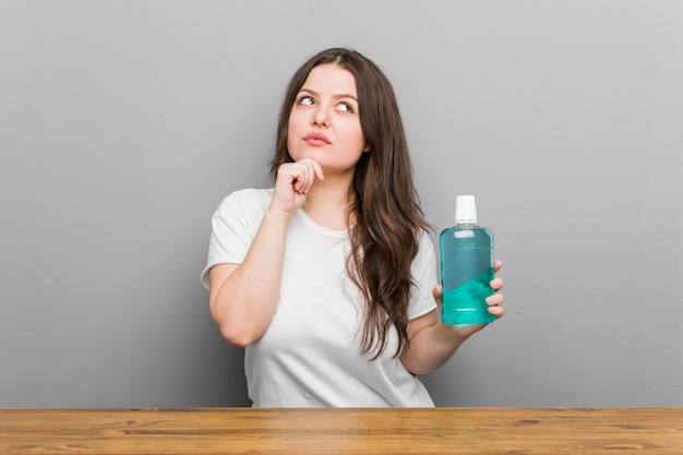 Jeune femme sinueuse de taille plus tenant un rince-bouche à côté avec une expression douteuse et sceptique.