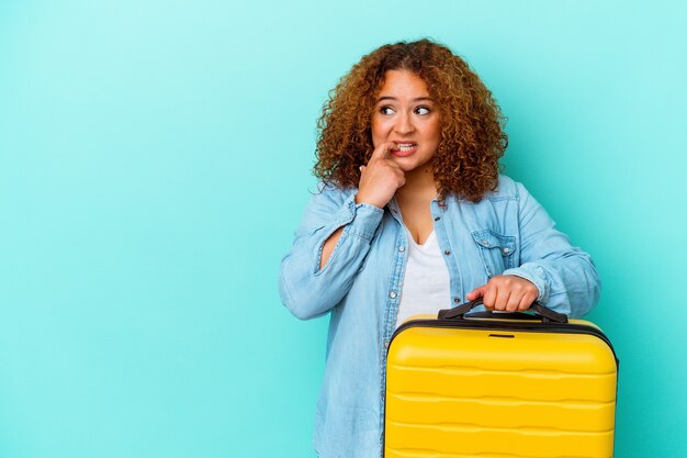 Jeune femme sinueuse de voyageur latin tenant une valise isolée sur fond bleu détendue en pensant à quelque chose en regardant un espace de copie.