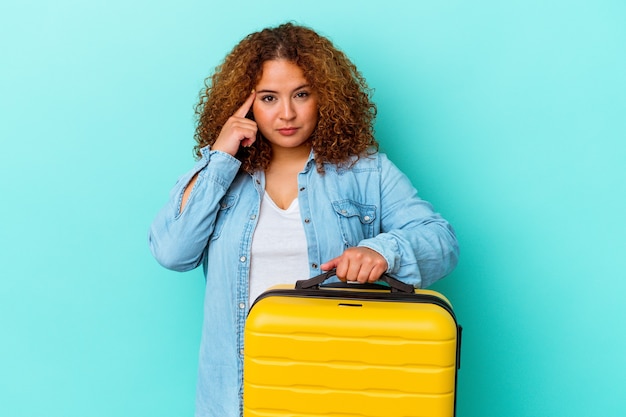 Jeune femme sinueuse de voyageur latin tenant une valise isolée sur fond bleu pointant le temple avec le doigt, pensant, concentré sur une tâche.