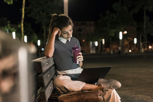 Photo jeune femme avec smoothie assis sur un banc la nuit à l'aide d'un ordinateur portable