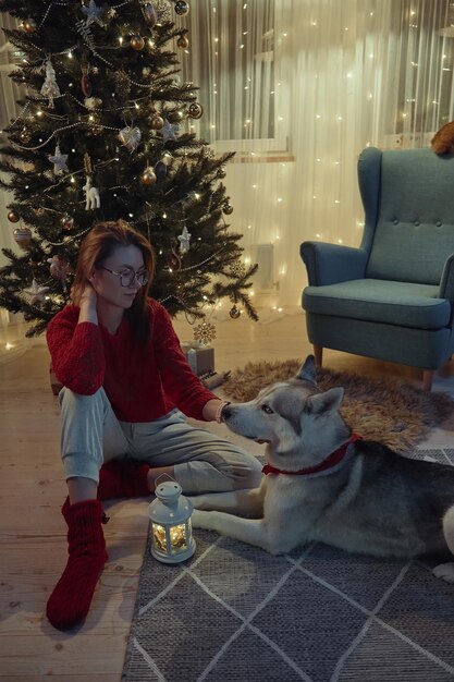 Jeune femme le soir de Noël avec ses animaux de compagnie caressant un chien assis sur le sol à côté de l'arbre de Noël