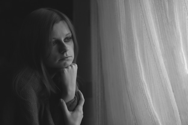 Jeune femme solitaire assise près de la fenêtre et regardant à travers elle