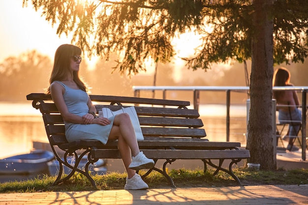 Jeune femme solitaire assise seule sur un banc au bord du lac profitant d'une chaude soirée d'été. Concept de bien-être et de détente dans la nature.