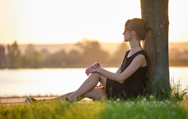 Jeune femme solitaire assise seule sur la pelouse au bord du lac profitant d'une soirée chaude Bien-être et détente dans le concept de la nature