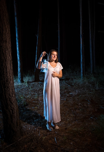 Jeune femme solitaire en chemise de nuit marchant dans la forêt la nuit avec lampe à gaz