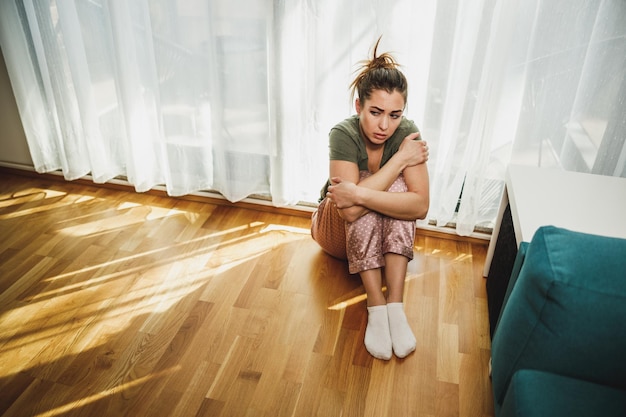 Une jeune femme solitaire en dépression s'assoit près de la fenêtre avec des genoux étreints perdus dans de mauvaises pensées pessimistes.