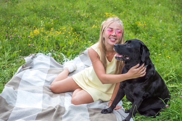 Jeune femme avec son gros chien noir est assise sur une couverture sur l'herbe dans une étreinte. Portrait de femme blonde à lunettes roses et chien