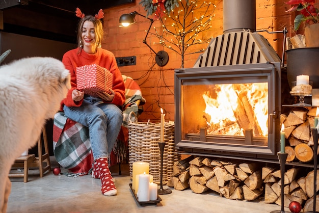 Jeune femme avec son mignon chien blanc pendant de joyeuses fêtes du Nouvel An assis près d'une cheminée à la maison