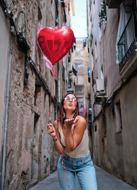 Jeune femme soufflant un baiser d'air à la caméra tout en tenant un ballon en forme de coeur rouge à l'extérieur dans la rue. Concept de l'amour et de la Saint-Valentin.