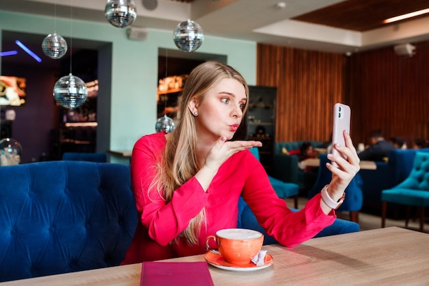 Jeune femme soufflant un baiser dans son smartphone communication en ligne médias sociaux à l'intérieur dans un café