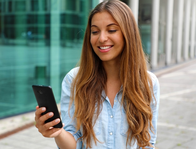 Jeune femme souriante à l'aide de son téléphone intelligent en se tenant debout devant un immeuble de bureaux