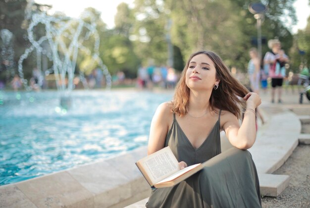 Une jeune femme souriante assise au bord d'une fontaine dans un parc lit un livre