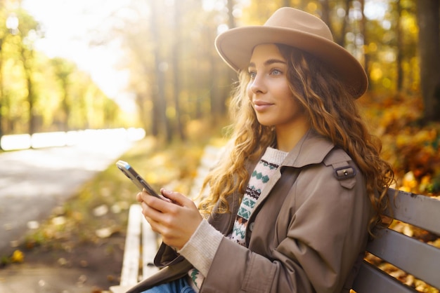 Une jeune femme souriante assise sur un banc dans un parc d'automne en utilisant un téléphone dans des vêtements élégants d'humeur heureuse