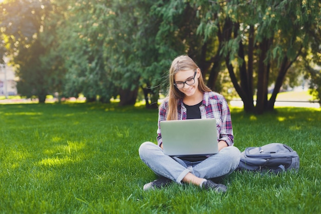 Jeune femme souriante assise à l'extérieur sur l'herbe avec ordinateur portable, tapant, surfant sur Internet, se préparant aux examens. Concept de technologie, de communication, d'éducation et de travail à distance, espace de copie