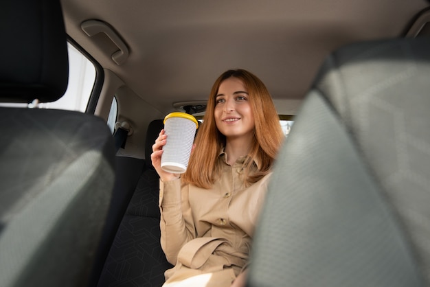 Jeune femme souriante assise sur le siège passager arrière et tenant une tasse de boisson chaude en attendant dans un embouteillage