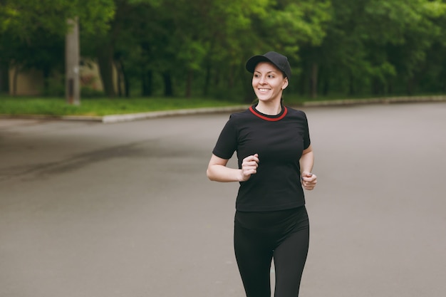 Jeune femme souriante athlétique belle brune en uniforme noir, entraînement à la casquette faisant des exercices de sport, course, jogging, regardant de côté sur le chemin dans le parc de la ville à l'extérieur