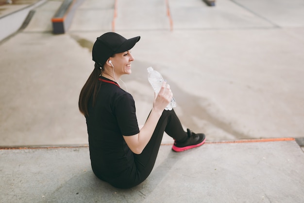Jeune femme souriante athlétique en uniforme noir, casquette avec casque écoutant de la musique tenant une bouteille d'eau assise avant ou après la course, entraînement dans un parc de la ville à l'extérieur