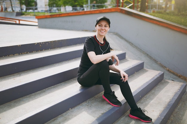 Jeune femme souriante athlétique en uniforme noir et casquette avec des écouteurs écoutant de la musique, se reposant et assise avant ou après avoir couru, s'entraînant dans les escaliers dans le parc de la ville à l'extérieur