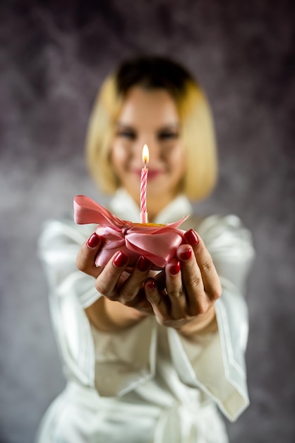 Une jeune femme souriante attrayante et excitée avec du maquillage et un peignoir tenant un gâteau d'anniversaire avec une bougie isolé sur un fond plat anniversaire en peignoire