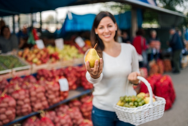 Jeune femme souriante au marché fermier. Tenant une poire à la main, regardant la caméra.