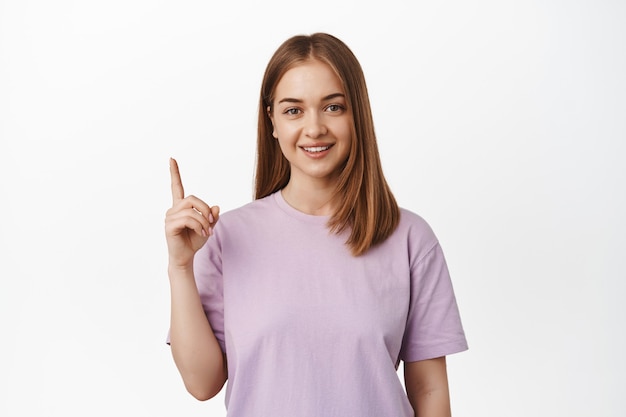 Jeune femme souriante aux cheveux blonds naturels, pointant le doigt vers le haut et l'air heureux, présente, fait une annonce, montre une bannière ou un logo, mur blanc