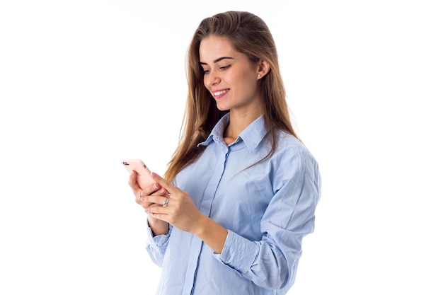 Jeune femme souriante aux cheveux longs en chemise bleue à l'aide de son smartphone rose en studio