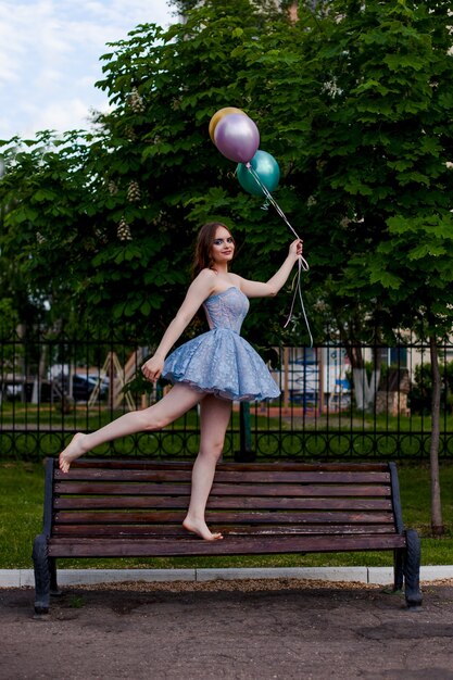 Une jeune femme souriante aux longues jambes vêtue d'une robe corset courte bleue marche pieds nus sur un banc et tient fl...