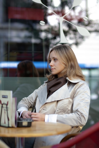 jeune femme souriante belle caucasienne portant un trench beige et une écharpe brune assise à la table