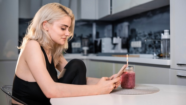 Jeune femme souriante blonde assise, utilisant son smartphone, une boisson saine