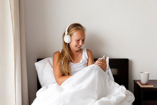 Jeune femme souriante blonde et heureuse écouter de la musique et boire du café dans un lit blanc le matin