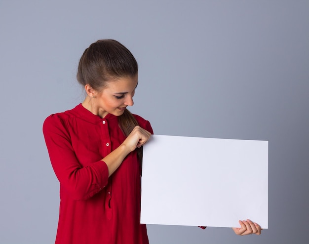 Jeune femme souriante en blouse rouge avec tresse montrant une feuille de papier blanche en studio