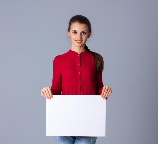 Jeune femme souriante en blouse rouge avec tresse montrant une feuille de papier blanche en studio
