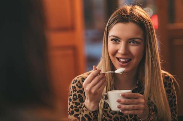 Une jeune femme souriante boit du café avec de la crème fouettée tout en étant assise au café avec un ami.