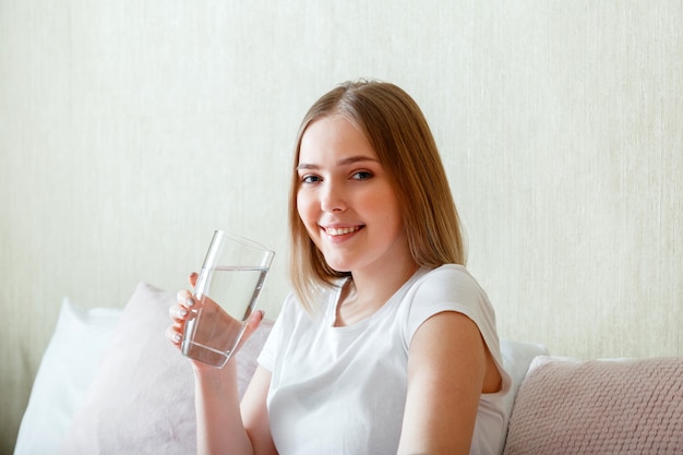 Une jeune femme souriante boit un verre d'eau pure le matin après s'être réveillée dans sa chambre. Une adolescente heureuse maintient l'équilibre de l'eau pour la santé du corps en buvant de l'eau propre.