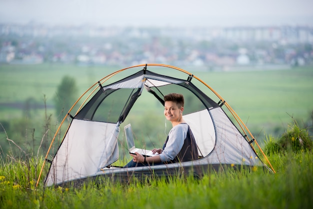 Jeune femme souriante à la caméra assise dans une tente au camping sur la colline en tapant sur son ordinateur portable