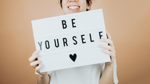 Jeune femme souriante à la caméra tout en tenant une pancarte soyez vous-même avec des vibrations positives à la caméra