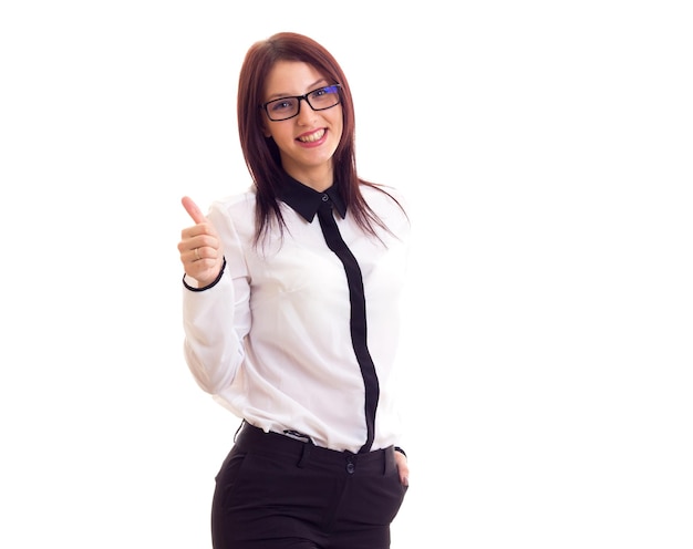 Jeune femme souriante en chemise blanche et pantalon noir avec de longs cheveux noirs et des lunettes noires montrant le pouce vers le haut sur fond blanc en studio