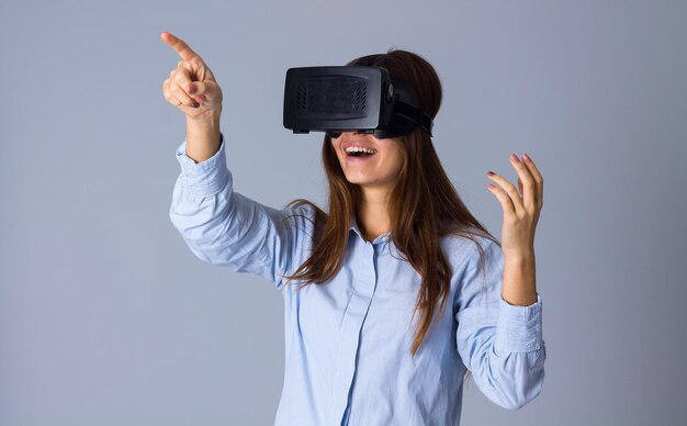 Jeune femme souriante en chemise bleue à l'aide de lunettes VR et pointant sur quelque chose en studio