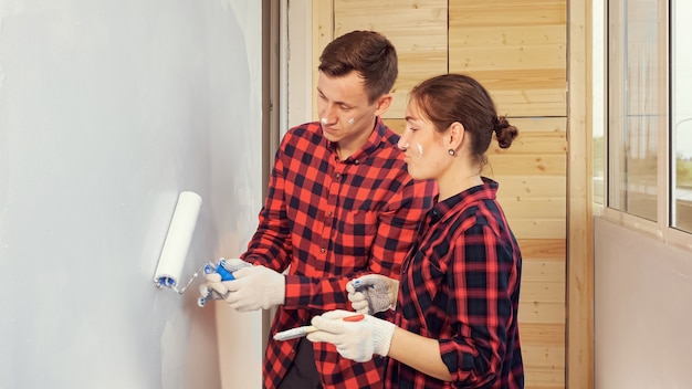 Une jeune femme souriante en chemise à carreaux rouge aide un mari joyeux à peindre un mur de couleur grise lors de la réparation sur une loggia vitrée à la maison