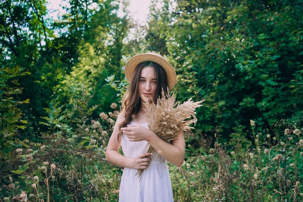 Jeune femme souriante dans un chapeau de paille sur fond de forêt verte d'été. Belle fille avec un bouquet de fleurs séchées lors d'une promenade. Fille dans une robe d'été.