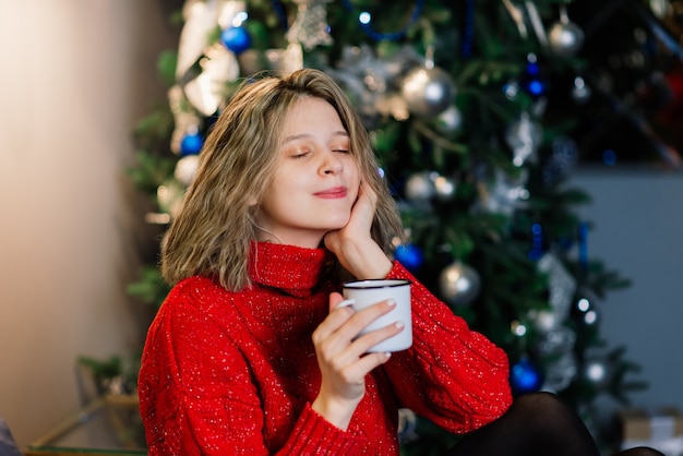Jeune femme souriante dans un pull, vacances d'hiver à l'intérieur de la maison décorée avec arbre de Noël.