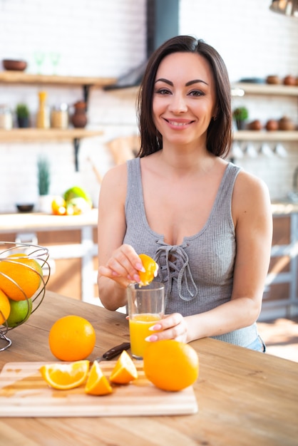 Une jeune femme souriante dans un t-shirt gris presse une orange fraîche sur du jus de fruits Concept d'alimentation saine À l'intérieur de la cuisine