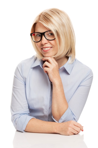Une jeune femme souriante dans des verres élégants et une chemise bleue est assise à la table. La réussite des entreprises. Isolé sur un mur blanc. Verticale.