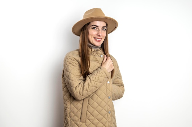 Jeune femme souriante dans une veste avec un chapeau contre un mur blanc