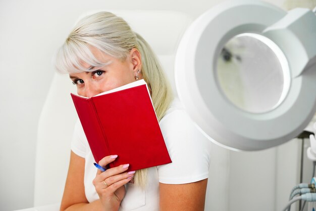 Une jeune femme souriante embarrassée avec un cahier rouge dans les mains est assise dans le bureau des esthéticiennes l...