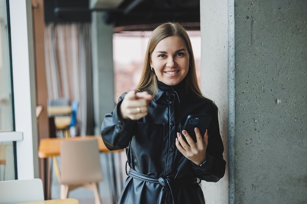 Une jeune femme souriante est debout avec un téléphone portable dans ses mains et regarde sur le côté tout en se tenant près d'une fenêtre reflétant la lumière du soleil Espace de travail moderne