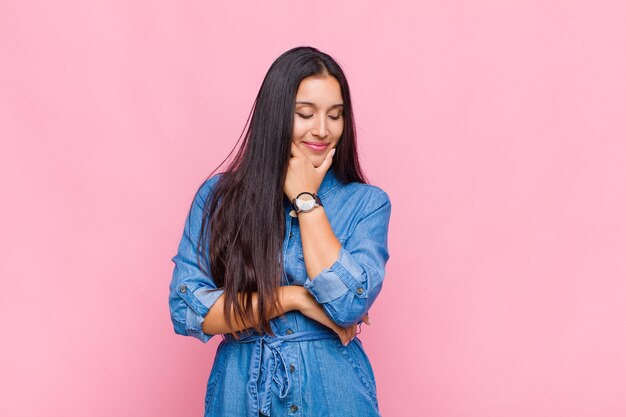 Jeune femme souriante avec une expression heureuse et confiante avec la main sur le menton, se demandant et regardant sur le côté