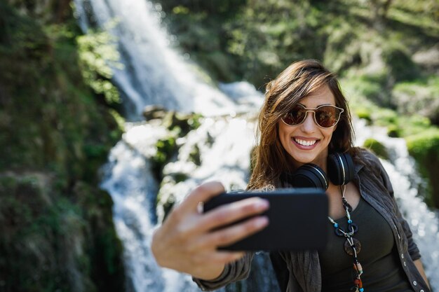 Une jeune femme souriante faisant du selfie avec son smartphone tout en profitant de la vue sur la magnifique cascade.