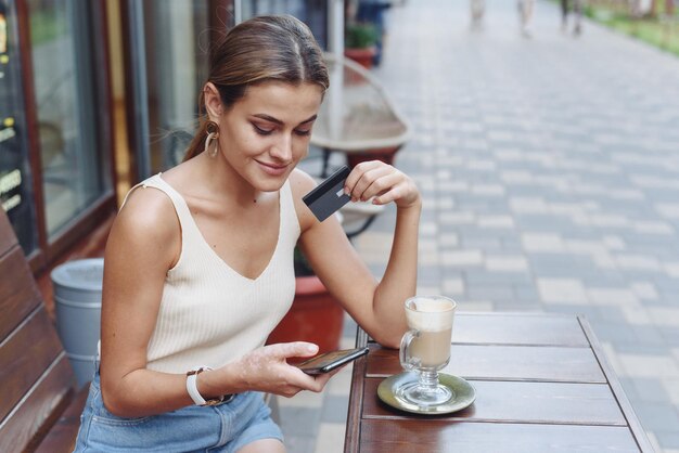 Jeune femme souriante faisant du shopping en ligne à l'aide d'un téléphone à l'extérieur dans un café Un beau modèle regarde le téléphone et boit du café en été